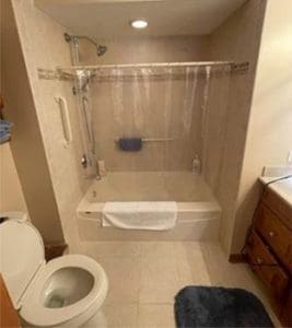 Small bathroom with a bathtub and shower, clear shower curtain, towel on the tub, toilet in the foreground, wooden vanity with sink and mirror to the right, and a blue bathmat on the floor.