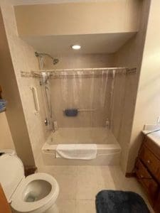 A bathroom with a white toilet, a bathtub with a clear shower curtain, beige tiled walls, a grab bar, a towel on the tub edge, a blue bath mat, and a wooden vanity with a sink.