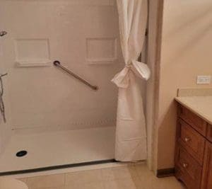 A bathroom with a walk-in shower featuring a grab bar, white curtain tied to the side, beige tile floor, and a wooden vanity with a light-colored countertop.