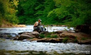 A man wearing outdoor gear sits on rocks by a flowing river surrounded by lush green trees, appearing to fish or rest with equipment beside him. The scene is peaceful and natural.