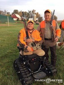 Two men wearing hunting gear and holding rifles pose outdoors on grass; one man is seated in a tracked wheelchair. A farm with barn, silo, and other buildings is visible in the background.