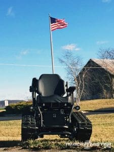 A tracked all-terrain wheelchair sits on grass under a clear blue sky, with a tall flagpole displaying the American flag waving in the background. A rustic barn and trees appear to the right.