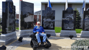 A man in a blue sweatshirt and white cap sits in a tracked wheelchair in front of a war memorial with engraved stone monuments and American flags in the background.