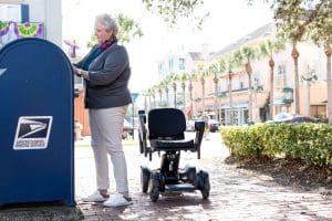 An older woman stands beside a USPS mailbox on a sunny sidewalk, mailing a letter. An empty mobility scooter is parked nearby. Shops and palm trees line the street in the background.