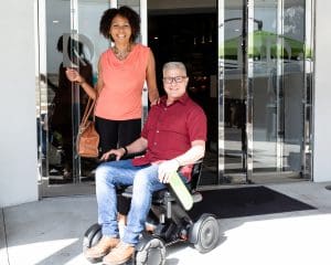 A smiling woman stands next to a smiling man in a wheelchair outside a building with glass doors. The man holds a green water bottle, and both are casually dressed.
