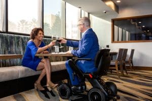 A woman and a man in a powered wheelchair sit at a restaurant table, smiling and toasting with glasses of wine. Both are dressed formally in blue attire, and sunlight shines through large windows behind them.