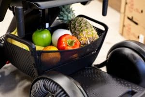 A close-up of a black mobility scooter basket filled with groceries, including a pineapple, green apple, red bell pepper, banana, orange, and a white onion. The scooter is parked indoors near cardboard boxes.