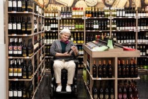 An older woman sits in a mobility scooter in a wine store aisle, examining a bottle of wine. Shelves filled with various bottles surround her on all sides.