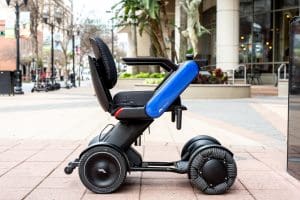 A modern, electric wheelchair with large wheels and a blue side panel is parked on a city sidewalk. Buildings, trees, and outdoor seating are visible in the background.