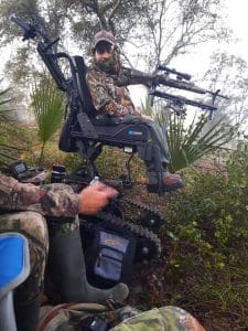 Person in camouflage gear sits in an all-terrain wheelchair with tank-like tracks, holding a crossbow in a wooded area, prepared for hunting. Another person in camouflage is partially visible nearby.