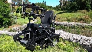 A black, tracked, all-terrain wheelchair is equipped with a mounted firearm on its front, positioned outdoors on grass with greenery and rocks in the background.