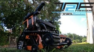 A rugged, black and orange all-terrain wheelchair with tank-like treads sits outdoors on grass. The sky and trees are in the background, along with the Track Freedom TrackMaster logo.