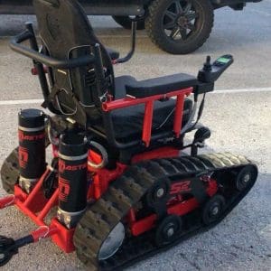 A motorized wheelchair with tank-like treads, red frame, and joystick controls is parked on asphalt, adapted for rugged terrain use. A black vehicle tire is visible in the background.