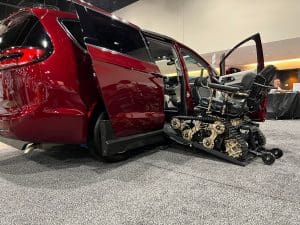A red minivan with its side door open features a specialized wheelchair with tank-like tracks, positioned on a ramp for easy access into the vehicle.
