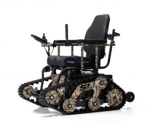 A camouflage-patterned all-terrain wheelchair with tank-like tracks, a padded seat, armrests, and control joystick, designed for mobility on rough surfaces.