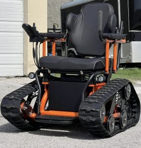 A black and orange all-terrain wheelchair with tank-style treads is parked outside on a paved surface near a building and trailer. The wheelchair has a padded seat, armrests with controls, and heavy-duty track wheels for rough terrain.