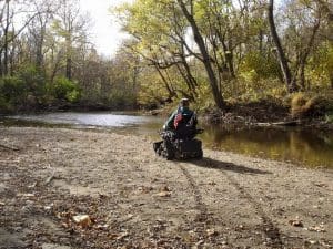 A person in a motorized wheelchair faces a calm river, surrounded by trees with autumn foliage on a sunny day. The ground is sandy and scattered with leaves and small branches.