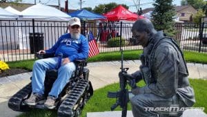 A man in a motorized wheelchair with tank-like treads sits beside a kneeling soldier statue holding a rifle. American flags and outdoor tents are visible in the background.