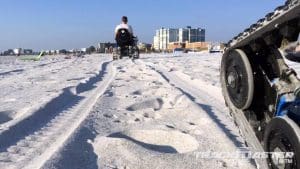 A person in a motorized wheelchair with tracks drives on a sandy beach, leaving deep tracks behind; a close-up of the wheelchairs tread is visible in the foreground. Buildings and blue sky are in the background.