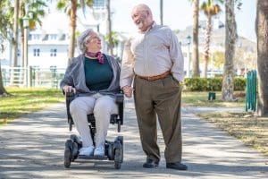 An elderly woman in a wheelchair and an elderly man walk hand in hand along a sunny, tree-lined path, smiling warmly at each other.
