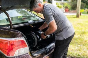 A man with gray hair places a lightweight mobility scooter into the trunk of a dark-colored car, outdoors on a sunny day with green grass and plants in the background.