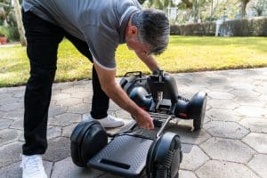 A man with gray hair in a gray polo shirt and black pants is assembling or adjusting a black go-kart or hoverboard on a paved outdoor pathway, with grass and trees in the background.