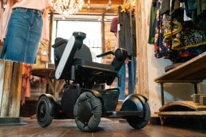 A modern electric wheelchair with large, rugged wheels is parked inside a boutique clothing store, surrounded by racks of clothes and wooden shelves on a wooden floor.