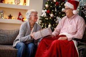 An elderly woman gives a wrapped gift to an elderly man in a Santa hat sitting in a wheelchair, both smiling, as festive decorations and a decorated Christmas tree highlight the spirit of Community Support Access.