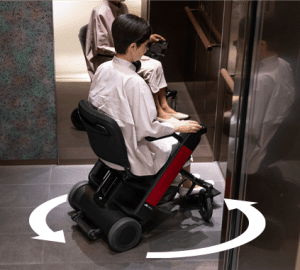 A person in a modern electric wheelchair rotates easily inside an elevator, demonstrating maneuverability with curved arrows indicating the turning motion.