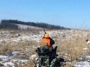 A person in an orange vest and hat is sitting on an all-terrain wheelchair, aiming a shotgun in a snowy, grassy field. Another person in orange is visible in the distance. The sky is clear and blue.