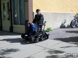 A man in a military uniform uses a tracked, all-terrain wheelchair on a sidewalk next to a yellow building. A motorcycle is partially visible in the background. The logo TRACKMASTER is at the bottom right.