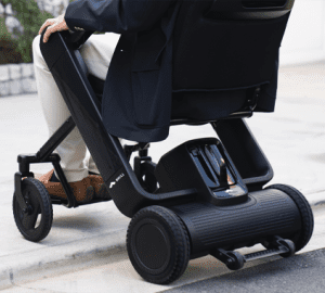 A person in light-colored pants and brown shoes uses a modern, black electric wheelchair to move over a curb outdoors. The wheelchair has large rear wheels and a sleek, compact design.