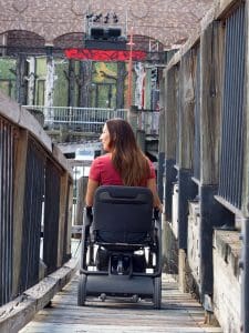 A woman with long brown hair in a red shirt uses a power wheelchair to travel across a narrow wooden ramp with railings, heading toward an outdoor stage area.