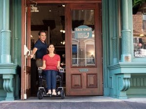 A smiling woman in a wheelchair is being pushed by a man as they exit a building through a wooden door with an Exit Only sign. The entrance is ornate with teal columns and trim.