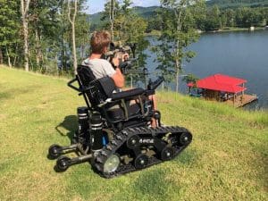 A person sits in an all-terrain, tank-like wheelchair on a grassy hill overlooking a lake with a red-roofed dock, holding a camera or binoculars. Trees and hills are visible in the background.