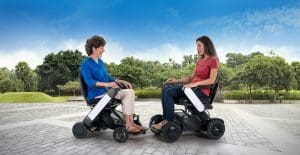 Two women sitting outdoors in electric wheelchairs face each other and smile, on a paved area with greenery and trees in the background under a blue sky.