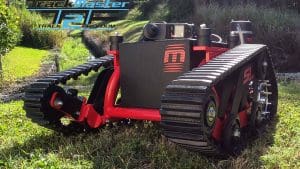 A red and black tracked robotic vehicle is positioned on grassy terrain with large rubber treads, cameras, and electronic equipment. TrackMaster T2F and logo are displayed in the upper left corner.