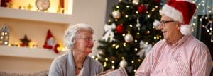 An elderly couple sits in front of a decorated Christmas tree, smiling at each other. The man wears a Santa hat and glasses; the woman holds a wrapped gift. Holiday decorations are visible in the background.