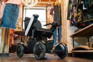 A modern electric wheelchair with large, sturdy wheels is positioned in the aisle of a cozy clothing boutique, surrounded by hanging garments and wooden shelves.