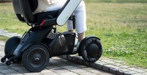 A person in white pants uses a modern electric wheelchair with large, rugged wheels to easily ascend a curb from a paved path onto the grass.