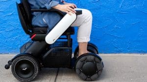 A person seated in a modern electric wheelchair with large, textured wheels, next to a bright blue textured wall. Only the persons lower body and hand are visible.