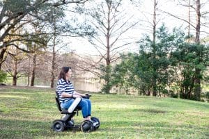 A woman in a striped shirt and jeans rides a motorized wheelchair across a grassy park area with trees and a pond in the background.