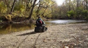 A person in a motorized wheelchair with an American flag drives along a sandy riverbank, surrounded by trees with autumn foliage, heading toward the water under a bright sky.