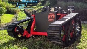 A rugged, red and black tracked robot vehicle sits on grass with a camera on top. The background shows a small ditch and greenery. The text TrackMaster TAF Track Freedom appears in the upper left corner.