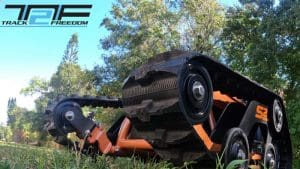 Close-up view of a rugged, orange and black all-terrain tracked mobility device labeled S2 on grass, surrounded by trees, with the Track Freedom logo in the corner.