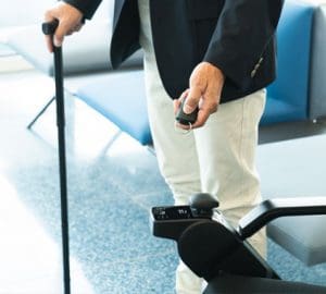 An older person holding a cane in their left hand and a remote for a wheelchair in their right hand.