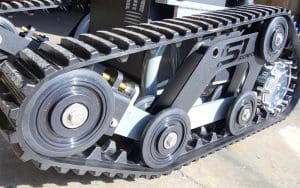 Close-up of a black rubber track system with metal gears and rollers, mounted on a machine or robot, shown on a concrete surface. The track features bolts and detailed mechanical components.