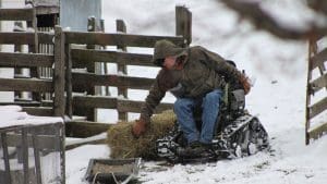 A person wearing a winter coat and gloves sits in an all-terrain wheelchair on snow-covered ground, moving a bale of hay near a wooden fence on a farm.