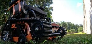 A close-up, low-angle view of a rugged, all-terrain wheelchair with tank-like treads, parked on grass with trees and a blue sky in the background.