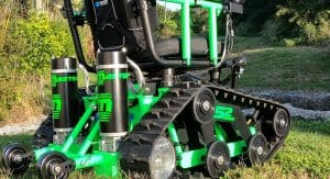 A close-up of a bright green, all-terrain tracked wheelchair with large rubber treads and suspension, positioned outdoors on grass near trees and foliage.